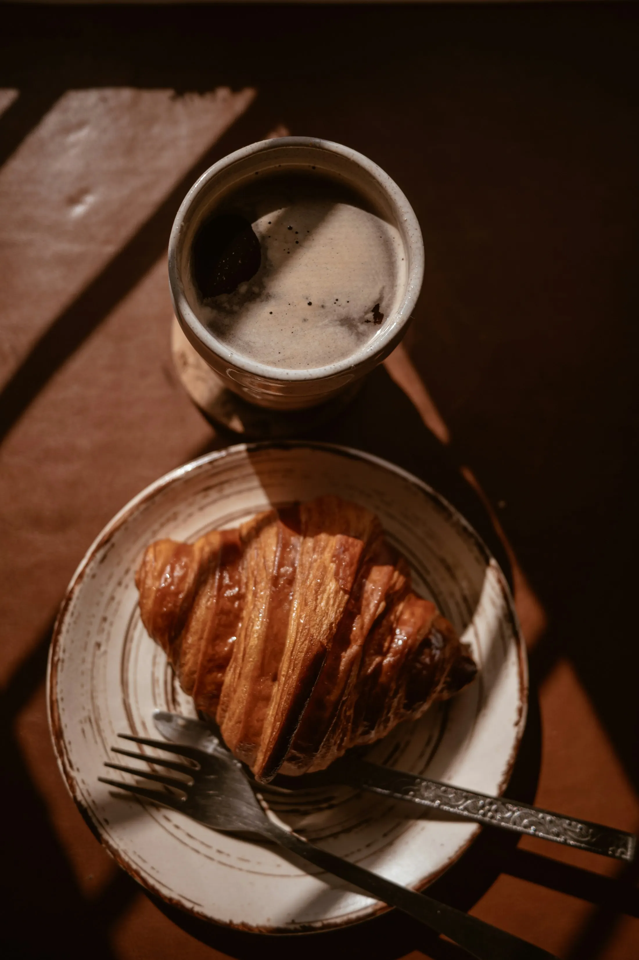 A croissant and a cup of coffee on a wooden table in warm morning light
