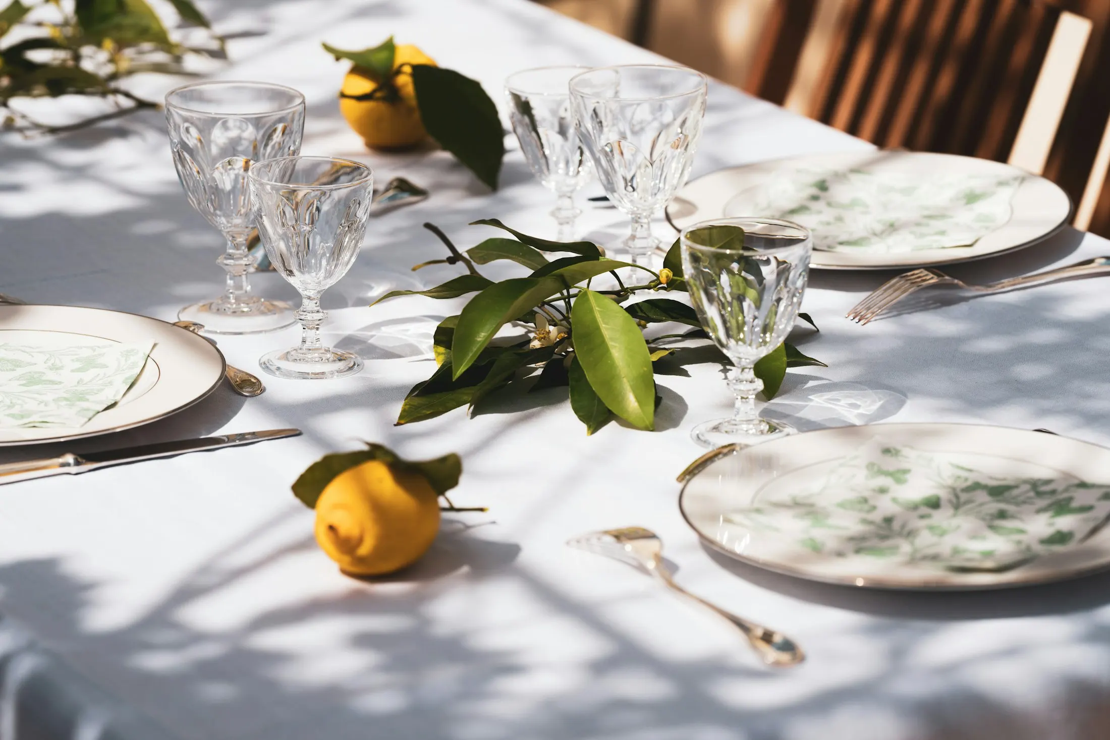 Lemons and glossy leaves on a white linen table set with glasses and plates, in dappled sunlight